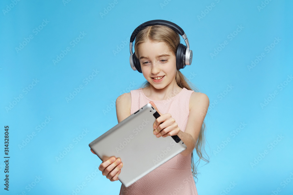 Beautiful little girl holding a tablet in her hands. The girl is wearing headphones. Portrait isolated on a blue background. Beautiful little girl holding a tablet in her hands. The girl is wearing headphones. Portrait isolated on a blue background.