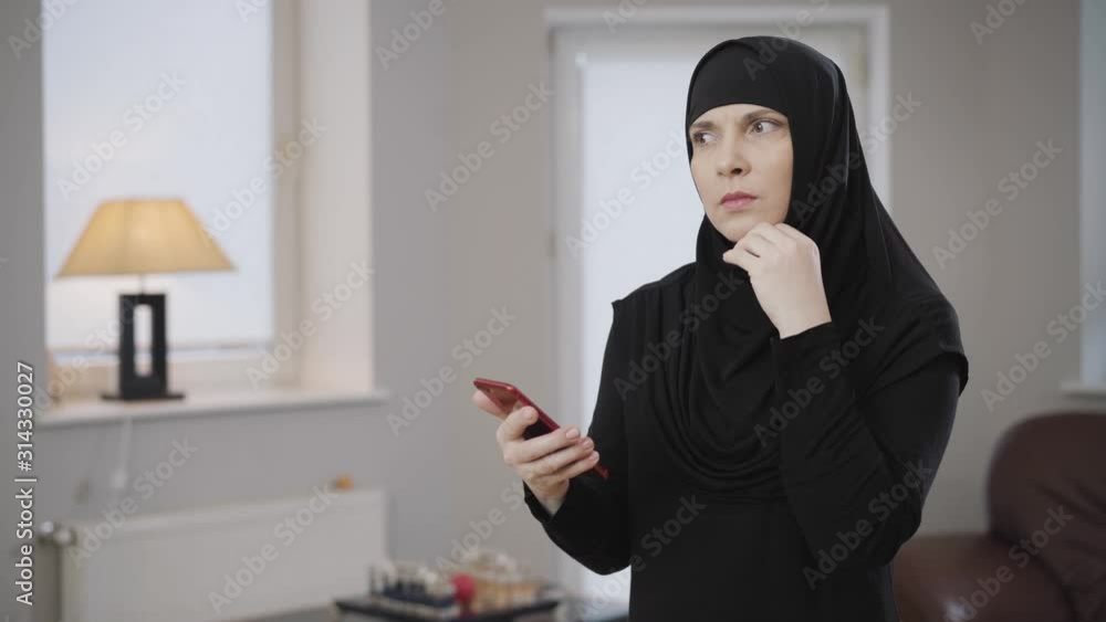 Side view portrait of young Muslim woman scrolling on smartphone and touching head with hand. Thoughtful eastern lady in black hijab using mobile phone. Eastern culture, modern society. Side view portrait of young Muslim woman scrolling on smartphone and touching head with hand. Thoughtful eastern lady in black hijab using mobile phone. Eastern culture, modern society.