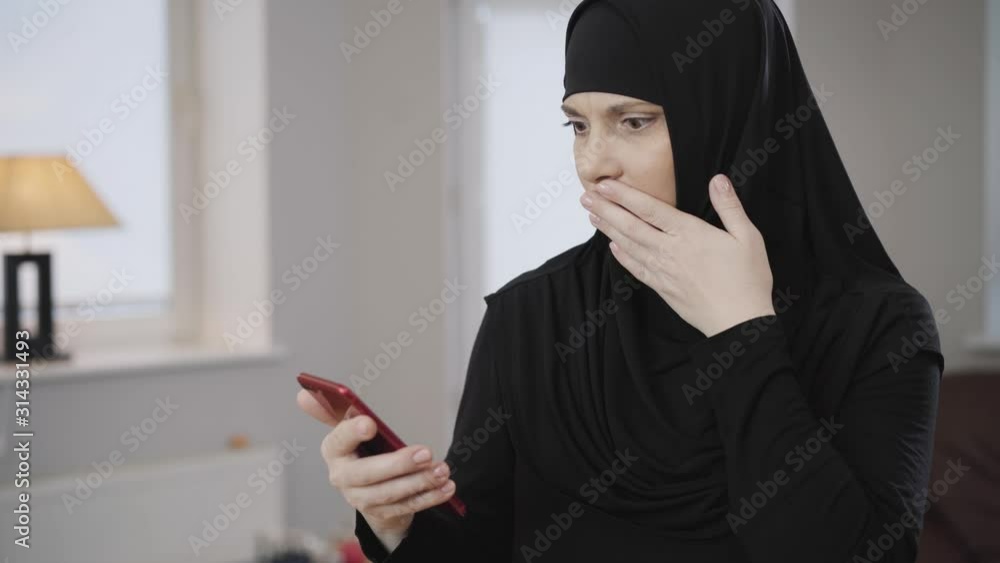 Young Muslim woman in traditional hijab looking at smartphone screen and making shocked facial expression. Eastern diligent lady shaking head. Traditional culture, modern society. Young Muslim woman in traditional hijab looking at smartphone screen and making shocked facial expression. Eastern diligent lady shaking head. Traditional culture, modern society.
