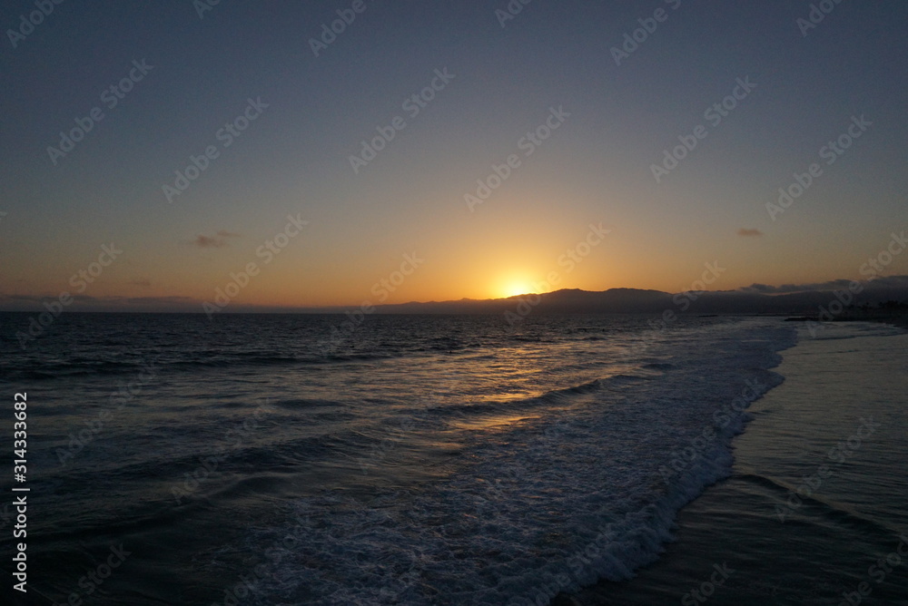 Sunsetting behind the mountains just off the beach of Santa Monica, California with a surfer and paraglider enjoying the views