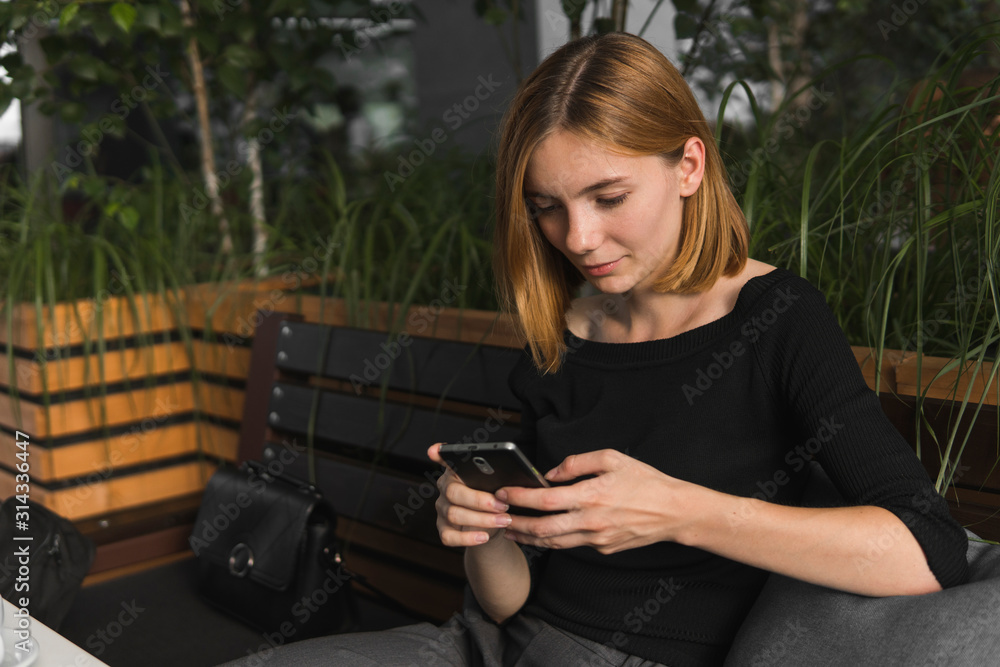 Young business woman uses phone. Good looking brunette female uses online banking on smart phone to transfer money from credit card. Girl using smart phone and chatting with business partners in cafe.