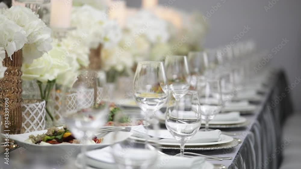 A festive table served by beautiful dishes in white colors surrounded by candles and peonies of flowers. Plates, cutlery and glasses for drinks. A festive table served by beautiful dishes in white colors surrounded by candles and peonies of flowers. Plates, cutlery and glasses for drinks.