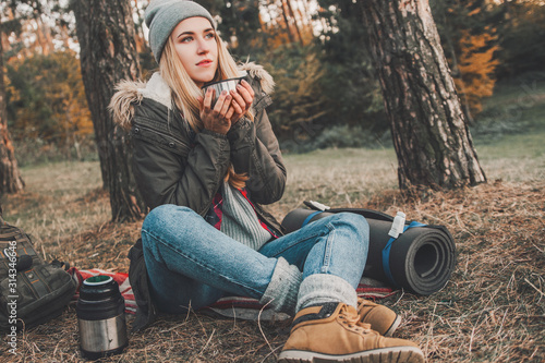 Hiking and travel along in the forest. Concept of trekking, adventure and seasonal vacation. Young woman with thermos sitting in woods. © zadorozhna