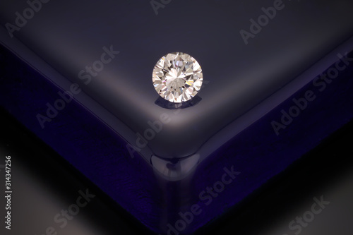 A solitaire, faceted, clear, round diamond sits on a reflective dark blue glass block background.