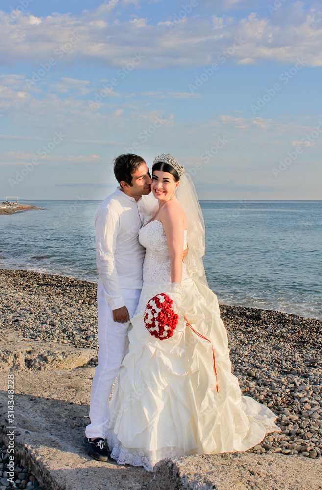 Wedding by the sea. Beautiful couple of newlyweds. Bride with a red bouquet in a wedding dress at the water. The groom kisses the bride on the cheek. Concept marriage, just married. Wedding by the sea. Beautiful couple of newlyweds. Bride with a red bouquet in a wedding dress at the water. The groom kisses the bride on the cheek. Concept marriage, just married.