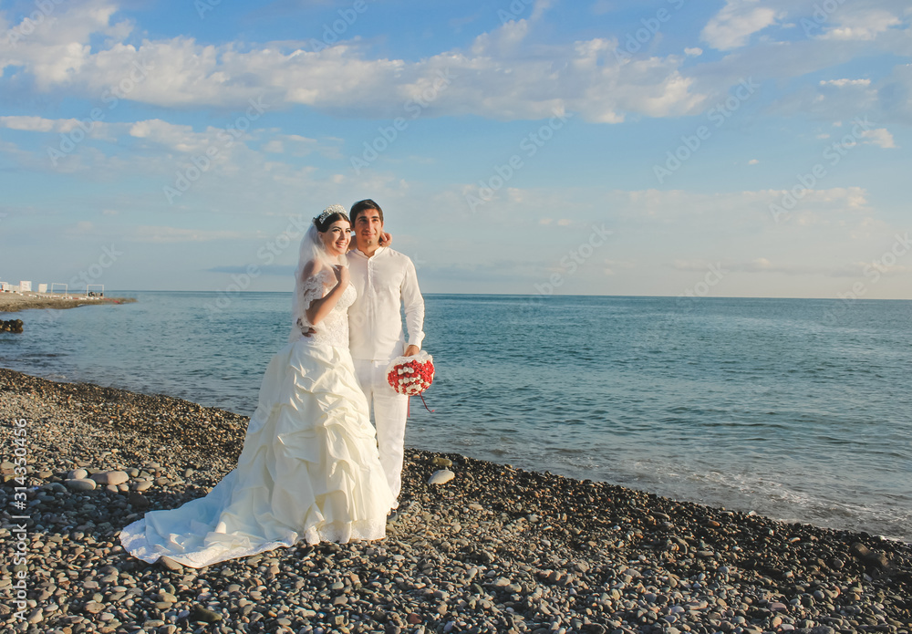 Wedding by the sea. Beautiful couple of newlyweds. Bride with a red bouquet in a wedding dress at the water. Bride and groom smile on stone beach during sunrise time. Just married in Batumi, Georgia. Wedding by the sea. Beautiful couple of newlyweds. Bride with a red bouquet in a wedding dress at the water. Bride and groom smile on stone beach during sunrise time. Just married in Batumi, Georgia.