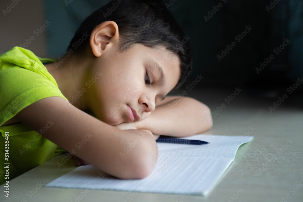 The child, the schoolboy does not want to do difficult homework, sits at the table, bored