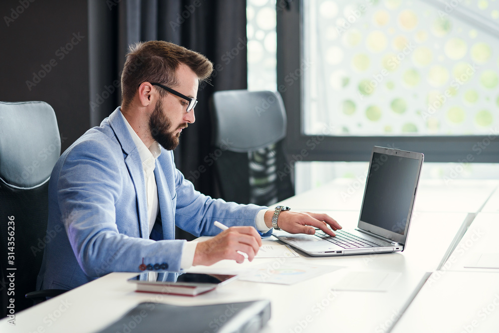 Successful bearded businessman in glasses and in blue suit works with laptop in modern office. Successful bearded businessman in glasses and in blue suit works with laptop in modern office.