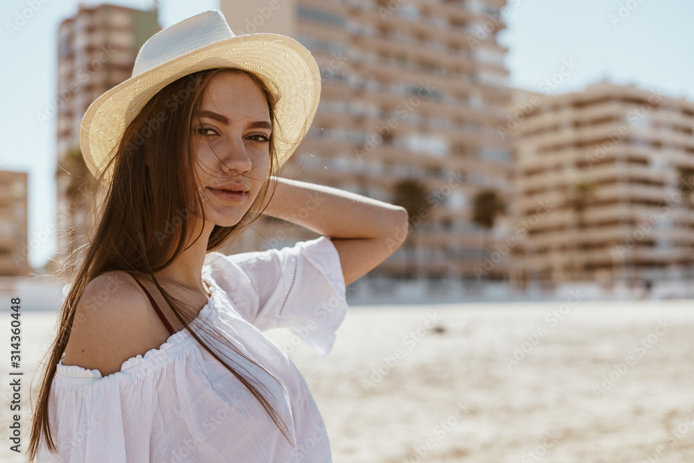 beautiful girl in a straw hat, a blouse on one shoulder attentively looks into the distance on the beach behind the house