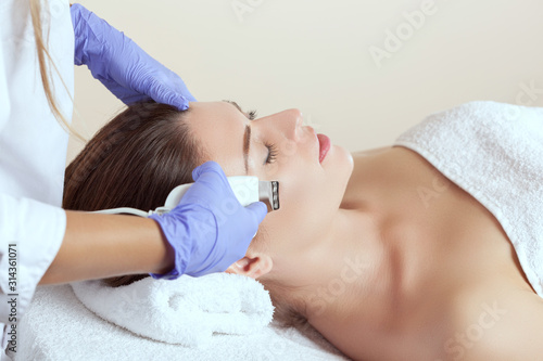 The doctor-cosmetologist makes the apparatus ultrasound cleaning procedure of of the facial skin of a beautiful young woman in a beauty salon. Cosmetology and professional skin care. photo