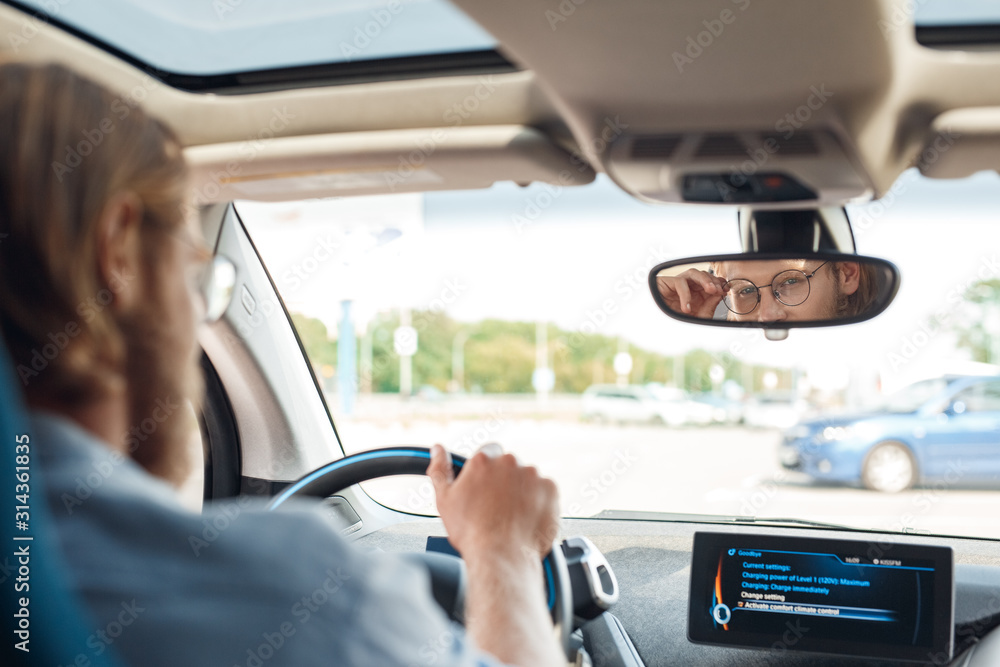 Transportation. Young man traveling by electric car sitting inside driving looking at rearview mirror touching eyeglasses playful back view