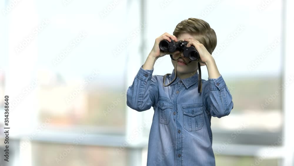Smiling caucasian boy looking through binoculars. Little boy in denim shirt using binoculars. Blurred window background. Smiling caucasian boy looking through binoculars. Little boy in denim shirt using binoculars. Blurred window background.