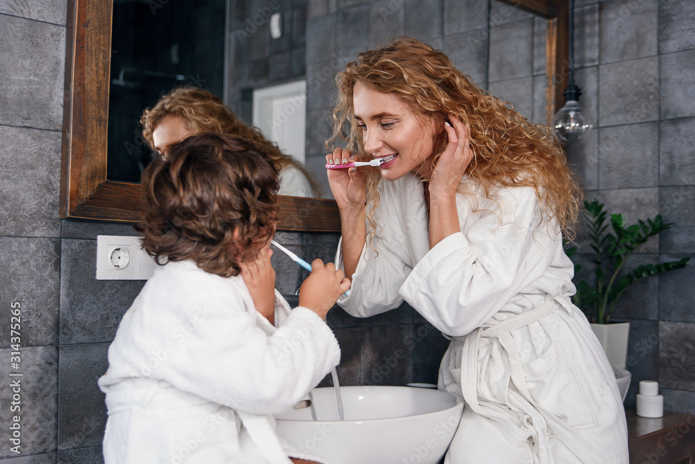 Young pretty woman with blonde hair teaches her little curly son to brush his teeth using a toothbrush and toothpaste. Young pretty woman with blonde hair teaches her little curly son to brush his teeth using a toothbrush and toothpaste.
