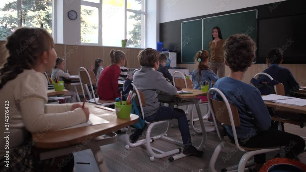 Focused elementary age children sitting at desks and listening to female teacher during art lesson in junior school. Diverse smart schoolkids hurrying to raise hands and answer teacher's question Focused elementary age children sitting at desks and listening to female teacher during art lesson in junior school. Diverse smart schoolkids hurrying to raise hands and answer teacher's question