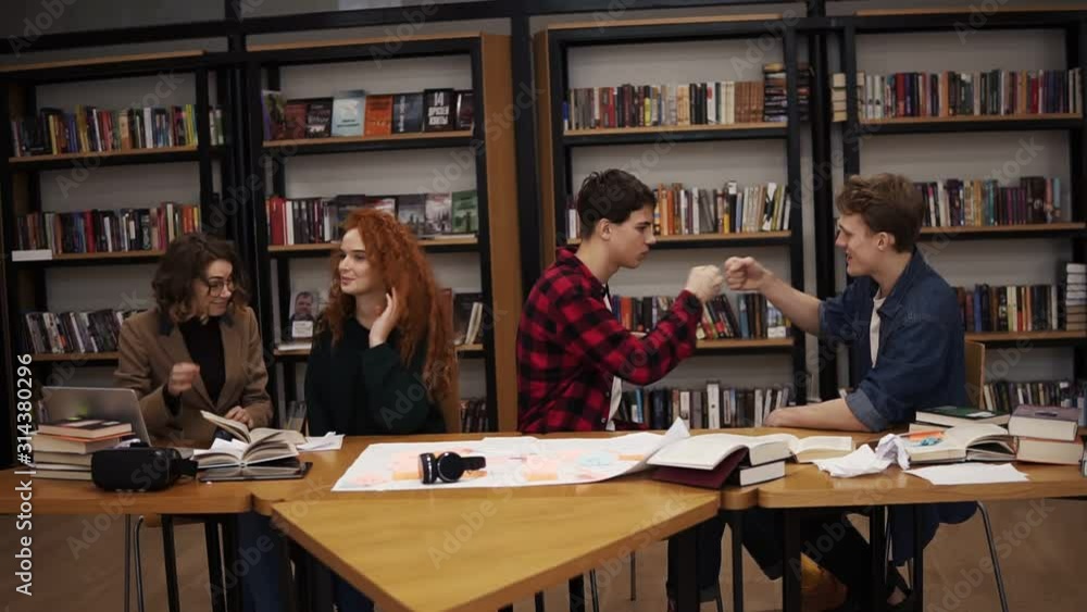 Slow motion of students making fun after hours of studying in uni library. Happy and free students - two men greeting each other, gesturing. Two girls discussing something sitting at the table in Slow motion of students making fun after hours of studying in uni library. Happy and free students - two men greeting each other, gesturing. Two girls discussing something sitting at the table in