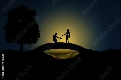 miniature people / toy photography - conceptual valentine holiday illustration. A man proposing a girl silhouette at the bridge under the moon light