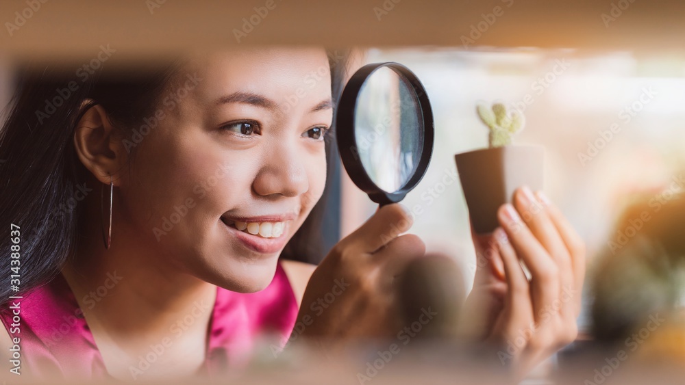 Asian smiling woman using a magnifying glass to look at the cactus in the planted pot.Concept idea of planting trees as a happy hobby. Asian smiling woman using a magnifying glass to look at the cactus in the planted pot.Concept idea of planting trees as a happy hobby.