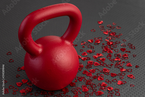 Fototapeta Naklejka Na Ścianę i Meble -  Holiday fitness, red kettlebell, with red heart confetti, on a black gym floor