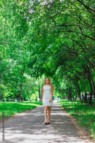 Portrait of a charming blond woman wearing beautiful white dress walking on the road under trees.