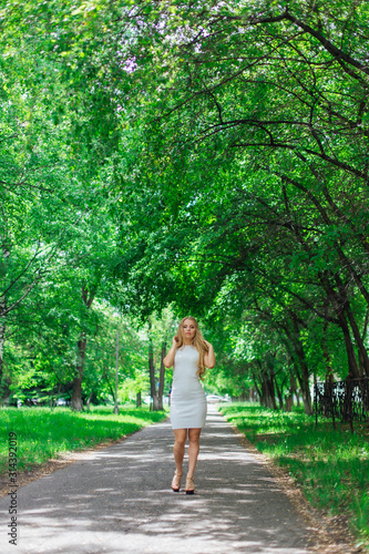 Portrait of a charming blond woman wearing beautiful white dress standing on the road under trees. © Smile