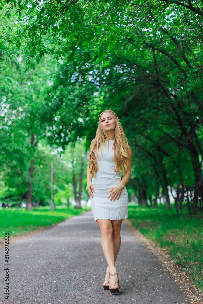 Portrait of a charming blond woman wearing beautiful white dress standing on the road under trees. Portrait of a charming blond woman wearing beautiful white dress standing on the road under trees.