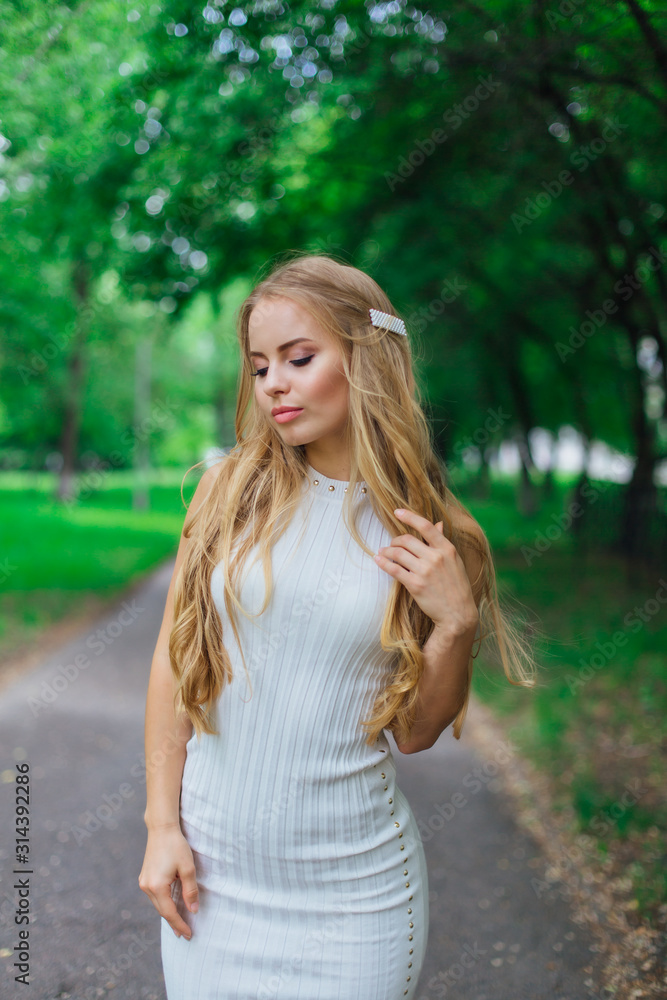 Portrait of a charming blond woman wearing beautiful white dress standing on the road under trees. Portrait of a charming blond woman wearing beautiful white dress standing on the road under trees.