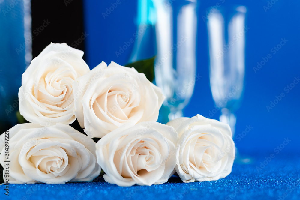 Obraz premium Two glasses of cold champagne and a bouquet of white roses on classic blue background. Saint Valentine's and women's day concept.