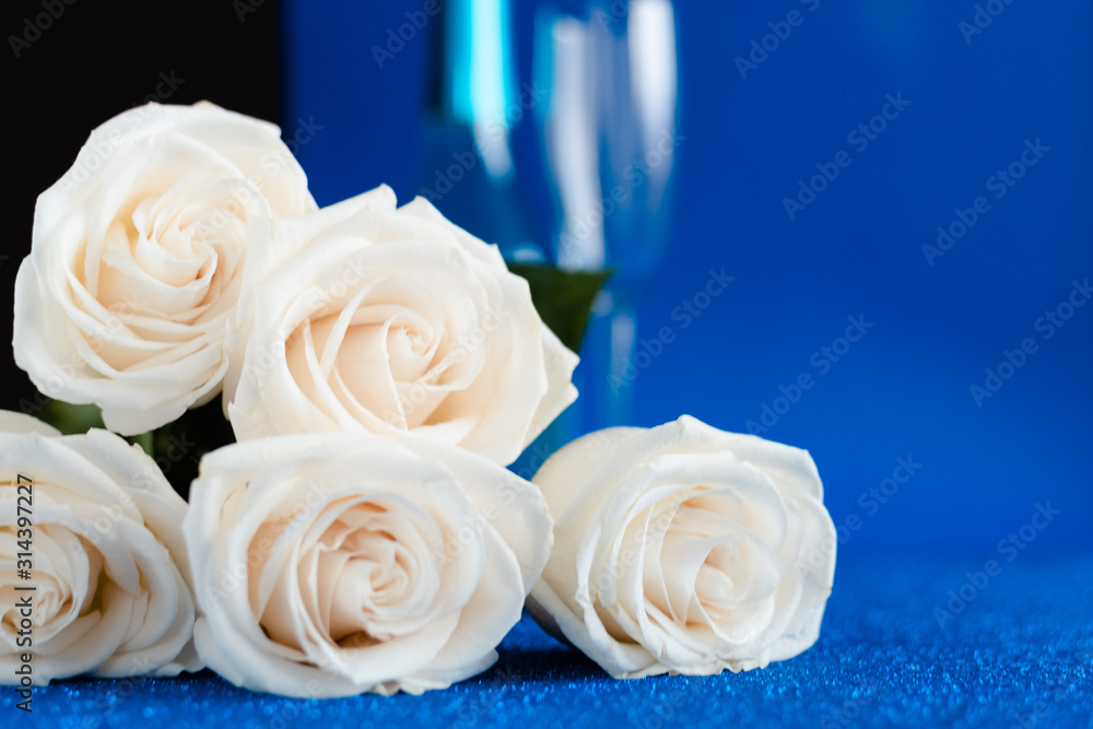 Fototapeta premium Two glasses of cold champagne and a bouquet of white roses on classic blue background. Saint Valentine's and women's day concept.