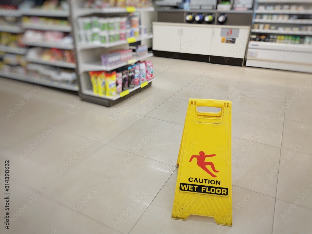 Yellow sign showing warning of caution wet floor. Wet floor sign is in the supermarket against the background of blurry products standing on the shelves. Selective focus. - obrazy, fototapety, plakaty 