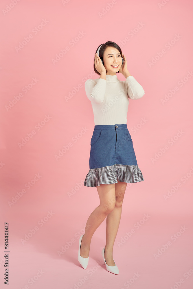 Full length portrait of a happy asian woman with headphones standing and holding mobile phone over pink background Full length portrait of a happy asian woman with headphones standing and holding mobile phone over pink background