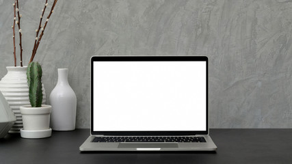 Cropped shot of modern workspace with blank screen laptop and ceramic vase on black desk with grey loft wall
