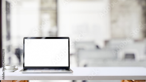 Cropped shot of simple workspace with blank screen laptop and coffee cup on white table with blurred office room