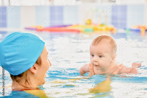 Young mother aged 30 years and baby son of 5 months are in pool. Smiling woman with beautiful snow-white smile is dressed in fashionable swimming suit swimsuit and cap. Girl teaches boy to swim.