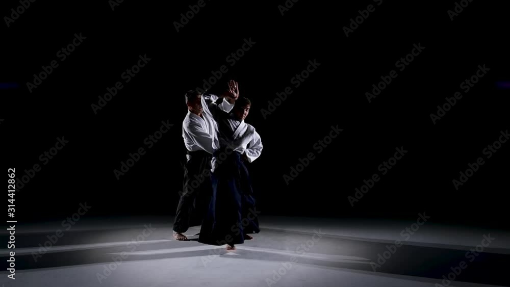 Two Masters participants of the training in special clothes of aikido hakama work out the methods of single combat on spotlights background. Slow motion. Two Masters participants of the training in special clothes of aikido hakama work out the methods of single combat on spotlights background. Slow motion.