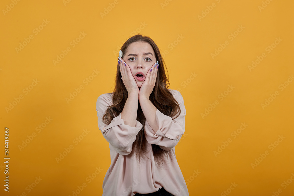 Surprised caucasian brunette woman in fashion pastel shirt keeping pals on cheeks, opened mouth, in shock on orange background in studio. People sincere emotions, lifestyle concept. Surprised caucasian brunette woman in fashion pastel shirt keeping pals on cheeks, opened mouth, in shock on orange background in studio. People sincere emotions, lifestyle concept.
