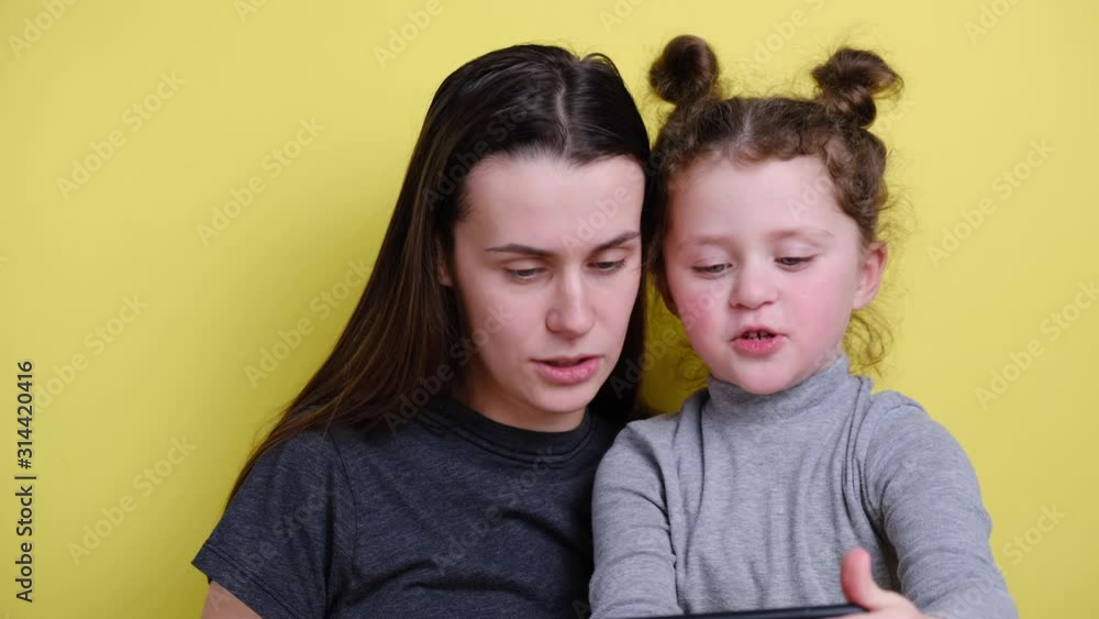 Happy young mum using social media app on smartphone recording video blog having fun with cute small kid girl embracing bonding together looking at mobile phone, sitting on background yellow wall Happy young mum using social media app on smartphone recording video blog having fun with cute small kid girl embracing bonding together looking at mobile phone, sitting on background yellow wall
