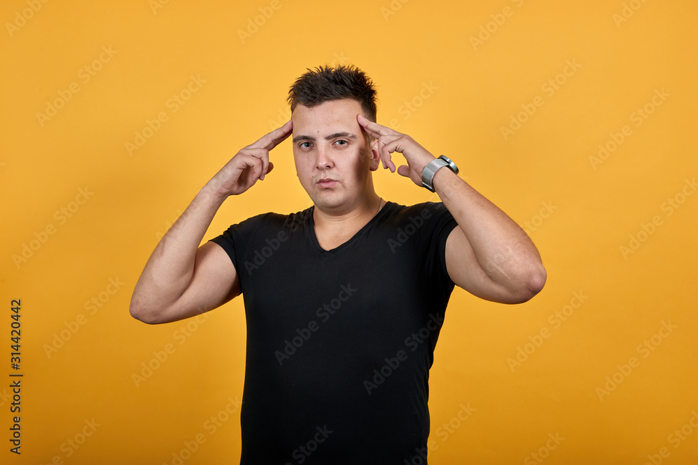 Focused caucasian young man keeping fingers on pulse, thinking, looking at camera wearing black shirt isolated on orange background in studio. People sincere emotions, lifestyle concept. Focused caucasian young man keeping fingers on pulse, thinking, looking at camera wearing black shirt isolated on orange background in studio. People sincere emotions, lifestyle concept.