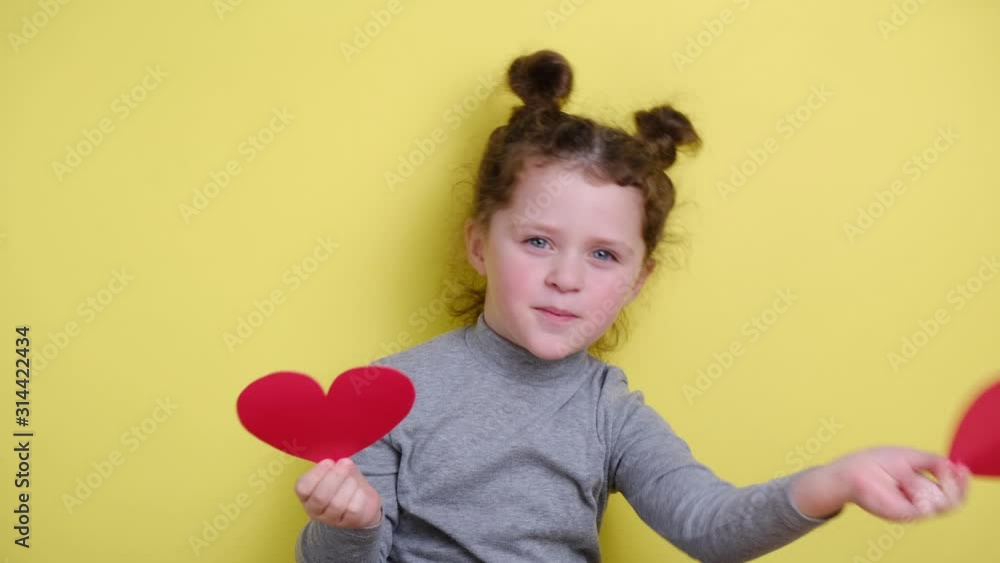 Funny little blogger looking at webcam, holding red hearts and smiling child girl talking to camera making video call vlog communicating online sitting on yellow background. Funny little blogger looking at webcam, holding red hearts and smiling child girl talking to camera making video call vlog communicating online sitting on yellow background.