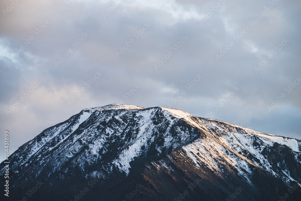 Atmospheric alpine landscape to snowy mountain ridge in sunset. Snow shines in golden light on mountain peak. Beautiful shiny snowy top. Evening cloudy sky. Wonderful scenery in gold sunset shades.