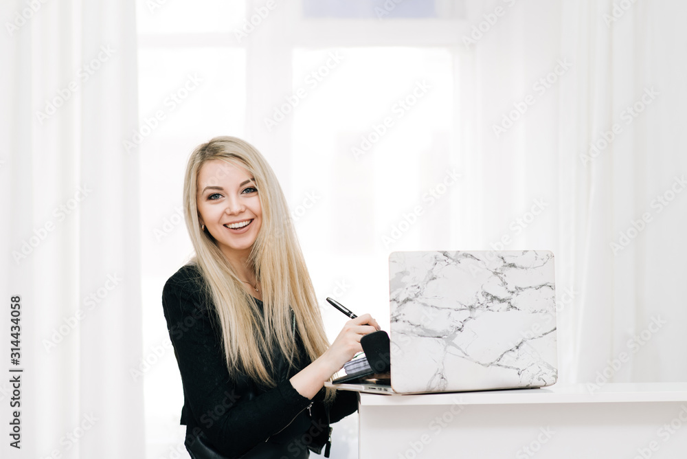 Beautiful smiling blonde girl with a notebook and pen in her hands, next to a laptop on a light background, office employee Beautiful smiling blonde girl with a notebook and pen in her hands, next to a laptop on a light background, office employee