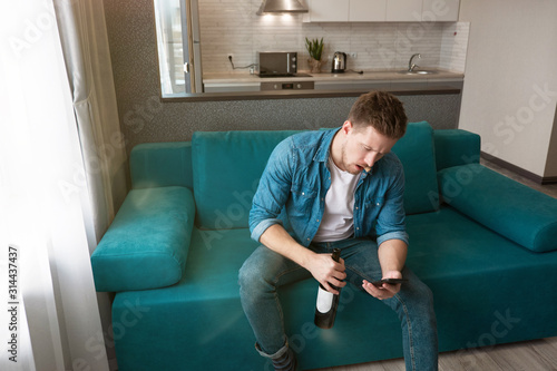 young handsome man sitting on the sofa with a bottle of wine texting message on his smartphone in the cozy living room home atmosphere photo