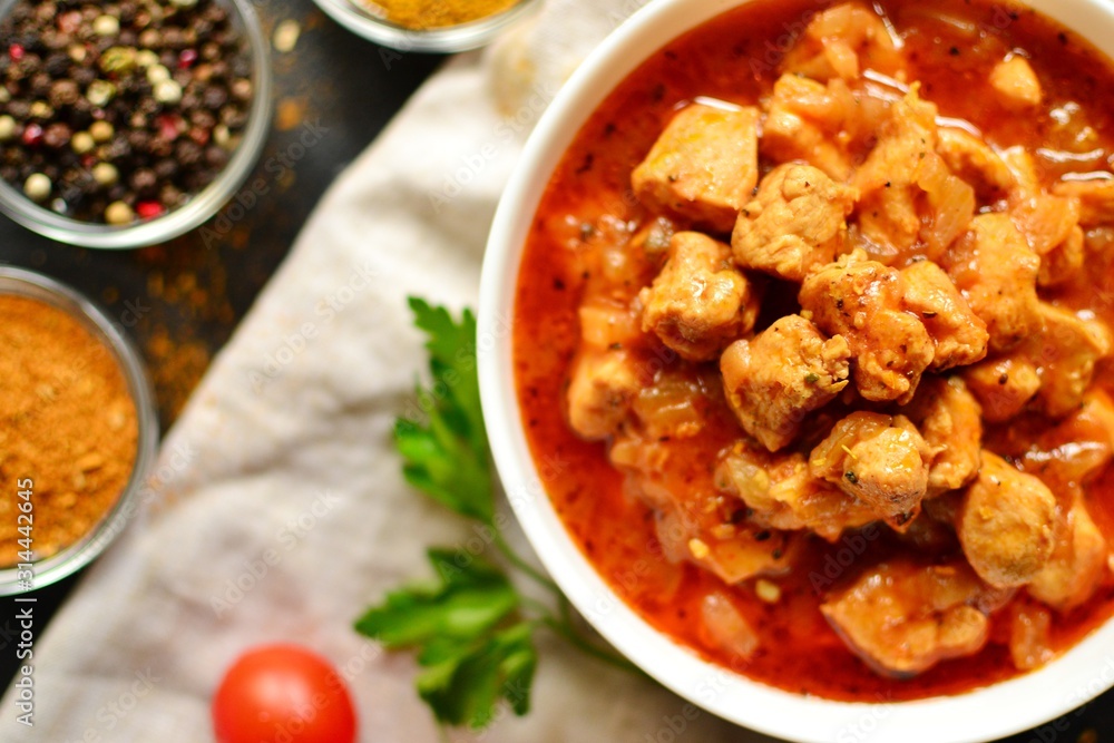 Tikka Masala. Chicken in tomato sauce in a white bowl. Spices and cherry tomatoes with parsley. Tasty chicken meat on a dark background. Horizontal view Tikka Masala. Chicken in tomato sauce in a white bowl. Spices and cherry tomatoes with parsley. Tasty chicken meat on a dark background. Horizontal view