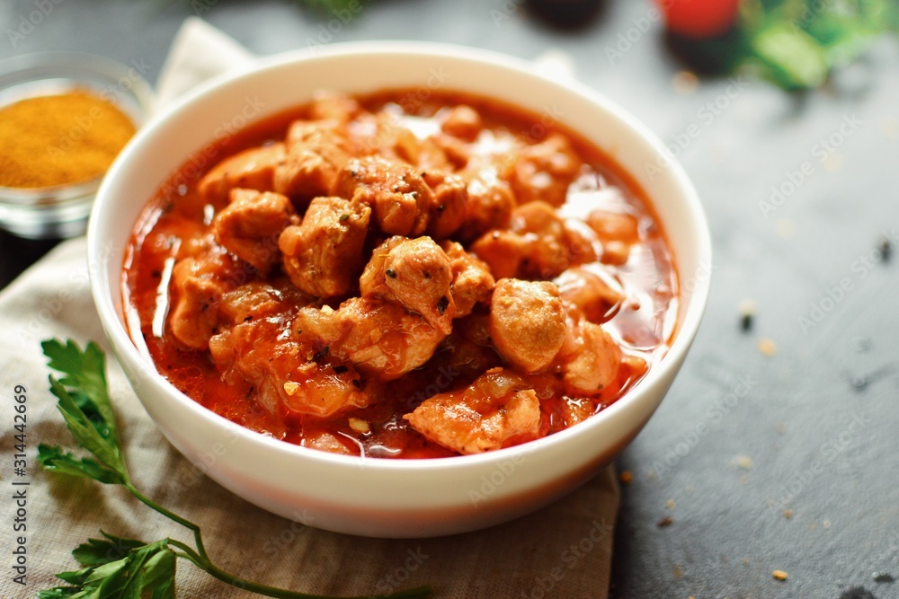Tikka Masala. Chicken in tomato sauce in a white bowl. Spices and cherry tomatoes with parsley. Tasty chicken meat on a dark background. Tikka Masala. Chicken in tomato sauce in a white bowl. Spices and cherry tomatoes with parsley. Tasty chicken meat on a dark background.