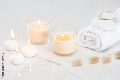 fluffy bunny tail grass near burning white candles in glass and rolled towel with stones on marble white surface