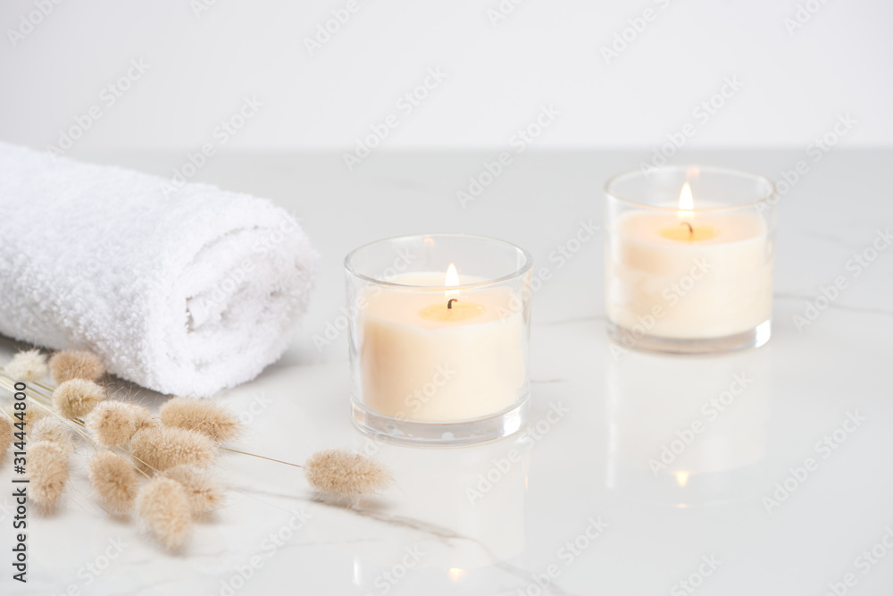 fluffy bunny tail grass near burning white candles in glass and rolled towel on marble white surface