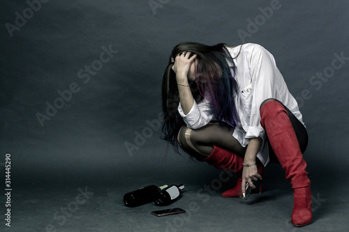 Brunette with dined hair in a white shirt and black shorts and red boots sits on a grey background and covers her face © Roman Kornev