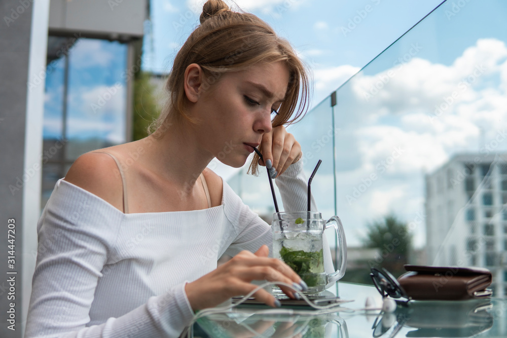 Cheerful blond woman blogger with cocktail is reading incoming sms message on smartphone connected to free wifi in cafe. Positive young student watching broadcasting online on modern mobile phone. Cheerful blond woman blogger with cocktail is reading incoming sms message on smartphone connected to free wifi in cafe. Positive young student watching broadcasting online on modern mobile phone.