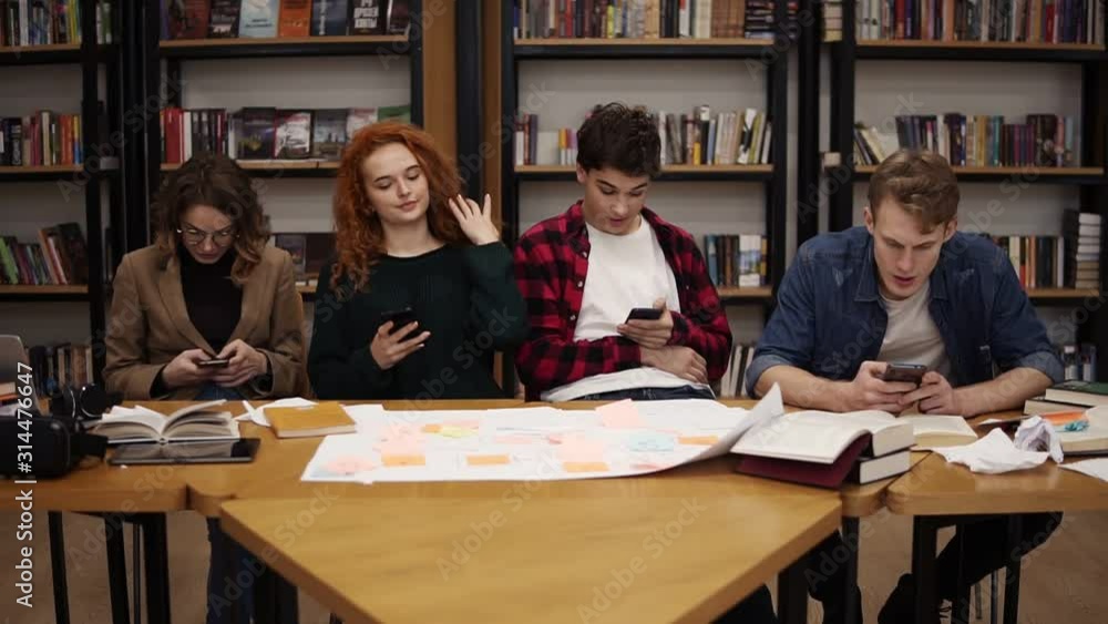 Group of four university students two males, two females sitting at the table together during break and using smartphones - texting, surfing internet while sitting in the uni library back to the Group of four university students two males, two females sitting at the table together during break and using smartphones - texting, surfing internet while sitting in the uni library back to the