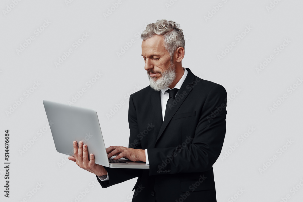 Confident mature man in full suit working using laptop while standing against grey background Confident mature man in full suit working using laptop while standing against grey background
