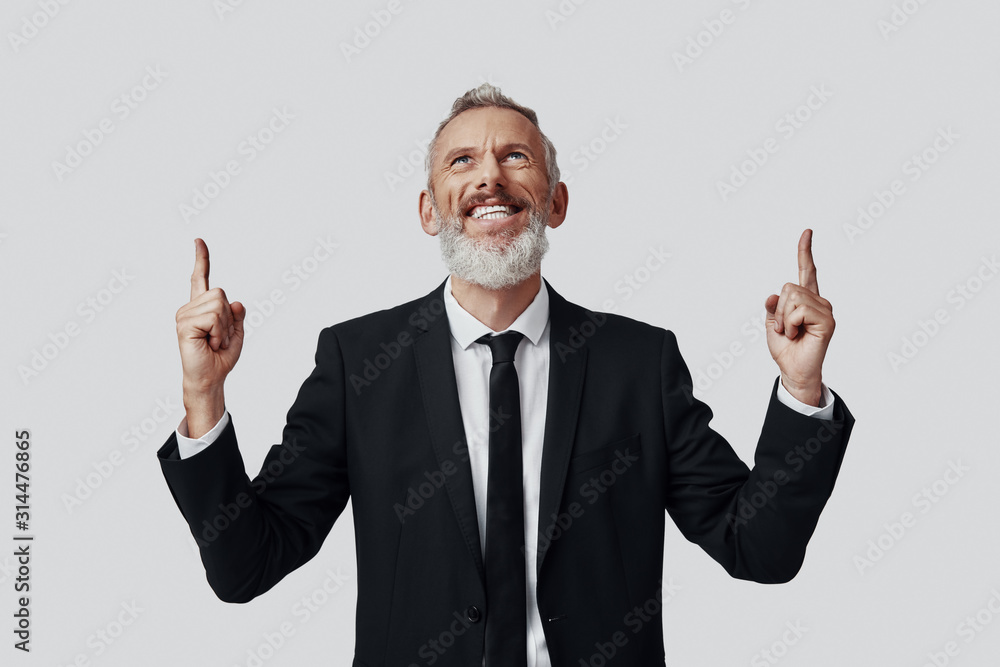 Happy mature man in full suit pointing copy space and smiling while standing against grey background Happy mature man in full suit pointing copy space and smiling while standing against grey background